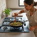 Mujer cocinando vegetales en sartén sobre parrilla de acero inoxidable de 5 quemadores empotrable en cocina moderna.