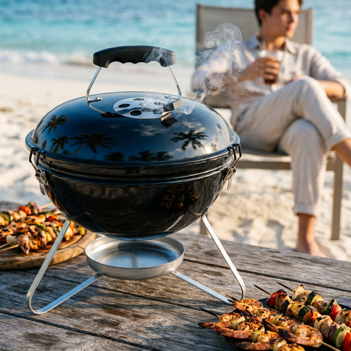 Asador de carbón portátil de 35 cm negro en una playa, humeando sobre una mesa de madera con brochetas de camarón y verduras.