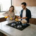 Pareja cocina en una isla moderna usando la Parrilla de Gas Avera de 4 quemadores y vidrio templado, con llamas azules activas.